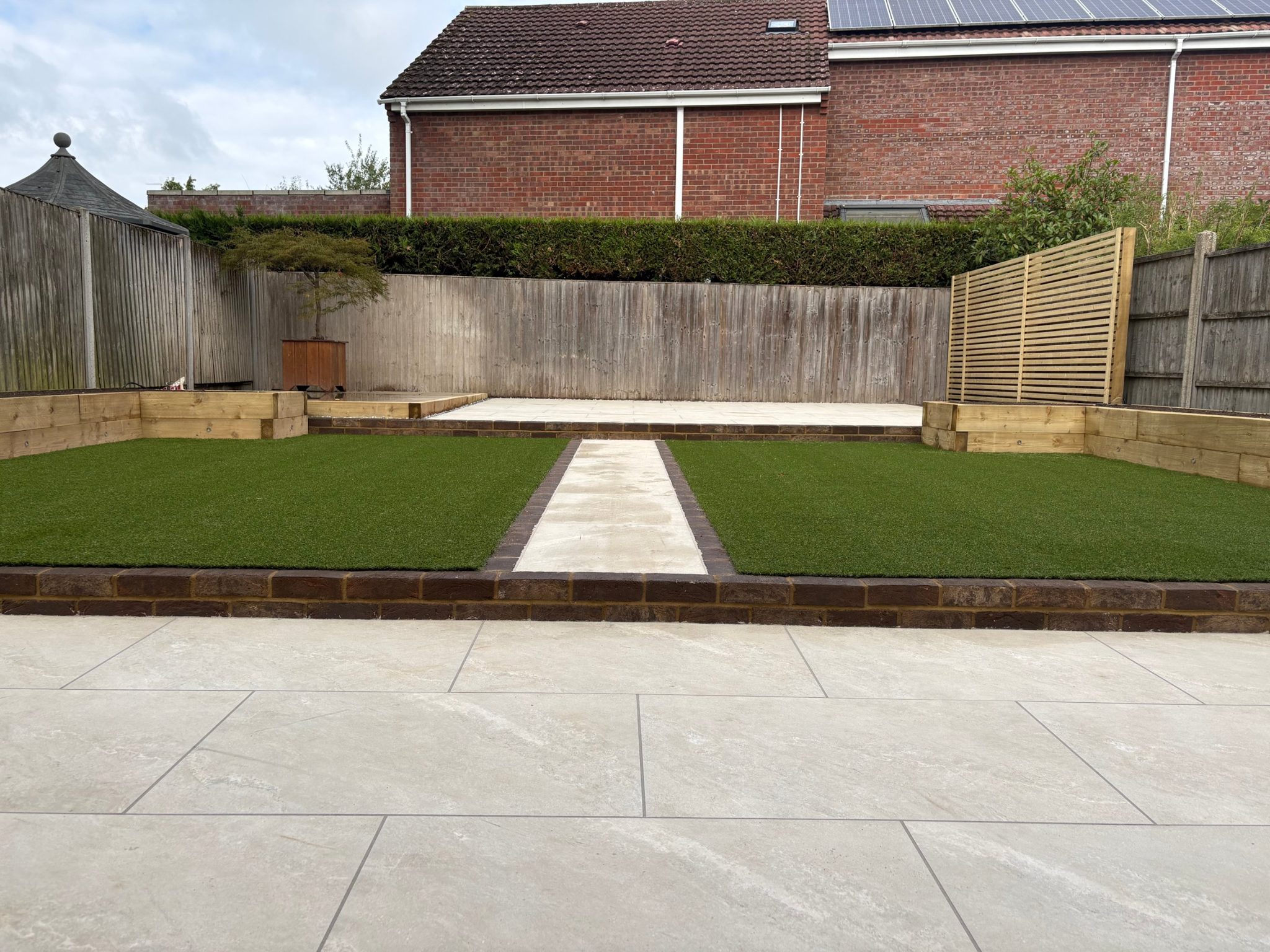 The middle tier: two artificial lawns either side of a brick-edged porcelain path, with sleeper raised beds wrapping both corners.