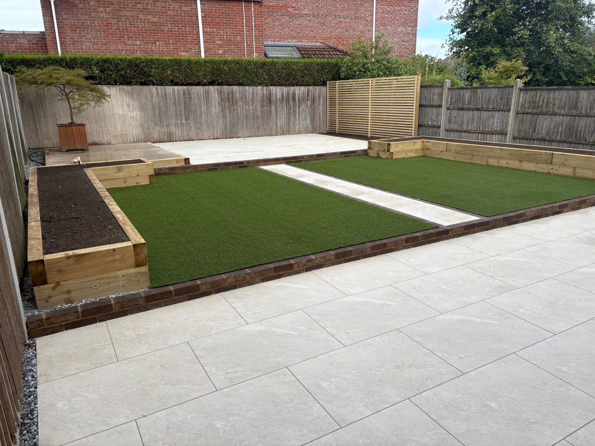 Across the middle tier from the corner of the terrace: dark brick step, two lawns, sleeper beds and the upper patio beyond.