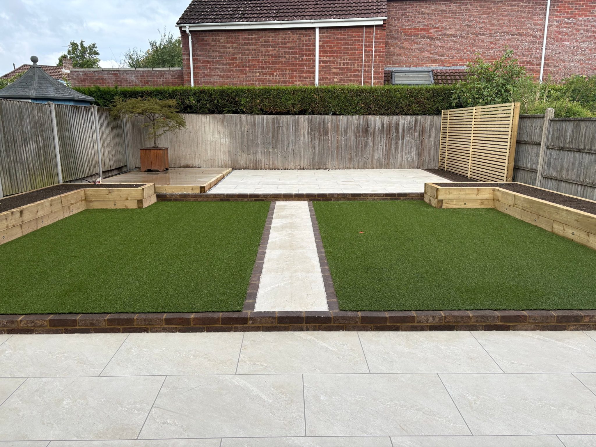 Three-tier garden in Fareham: Himalayan White porcelain terrace, a dark brick step up to two artificial lawns split by a porcelain path, sleeper raised beds and an upper porcelain patio