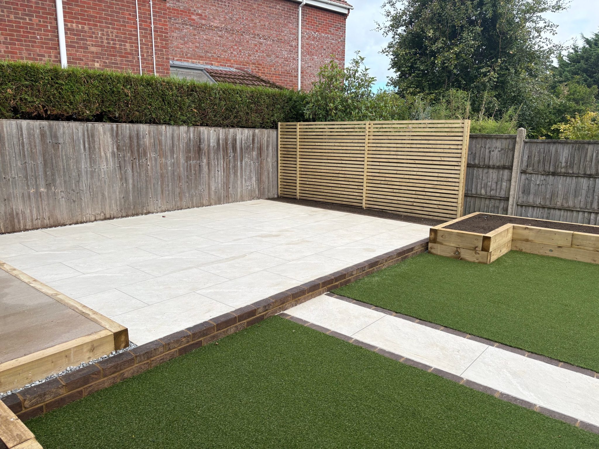 The upper tier: a raised Himalayan White porcelain patio framed in dark brick, with a Venetian-style slatted screen and a planting bed in the corner.