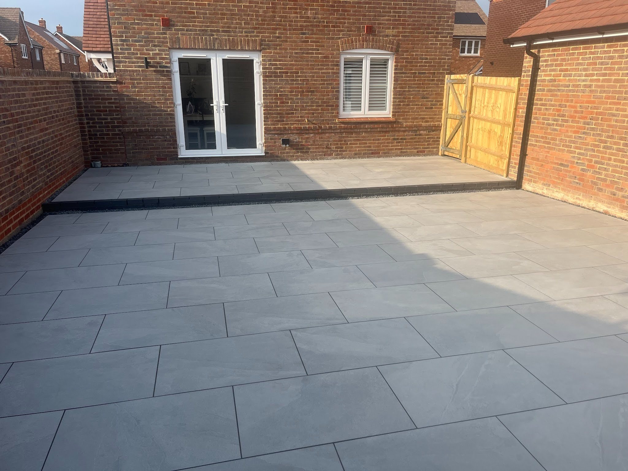 Wide view in dry conditions &mdash; the two-tier composition reads from ground level, both terraces in Victoria Grey porcelain with the dark engineering brick step and Derbyshire Peak gravel running between them.