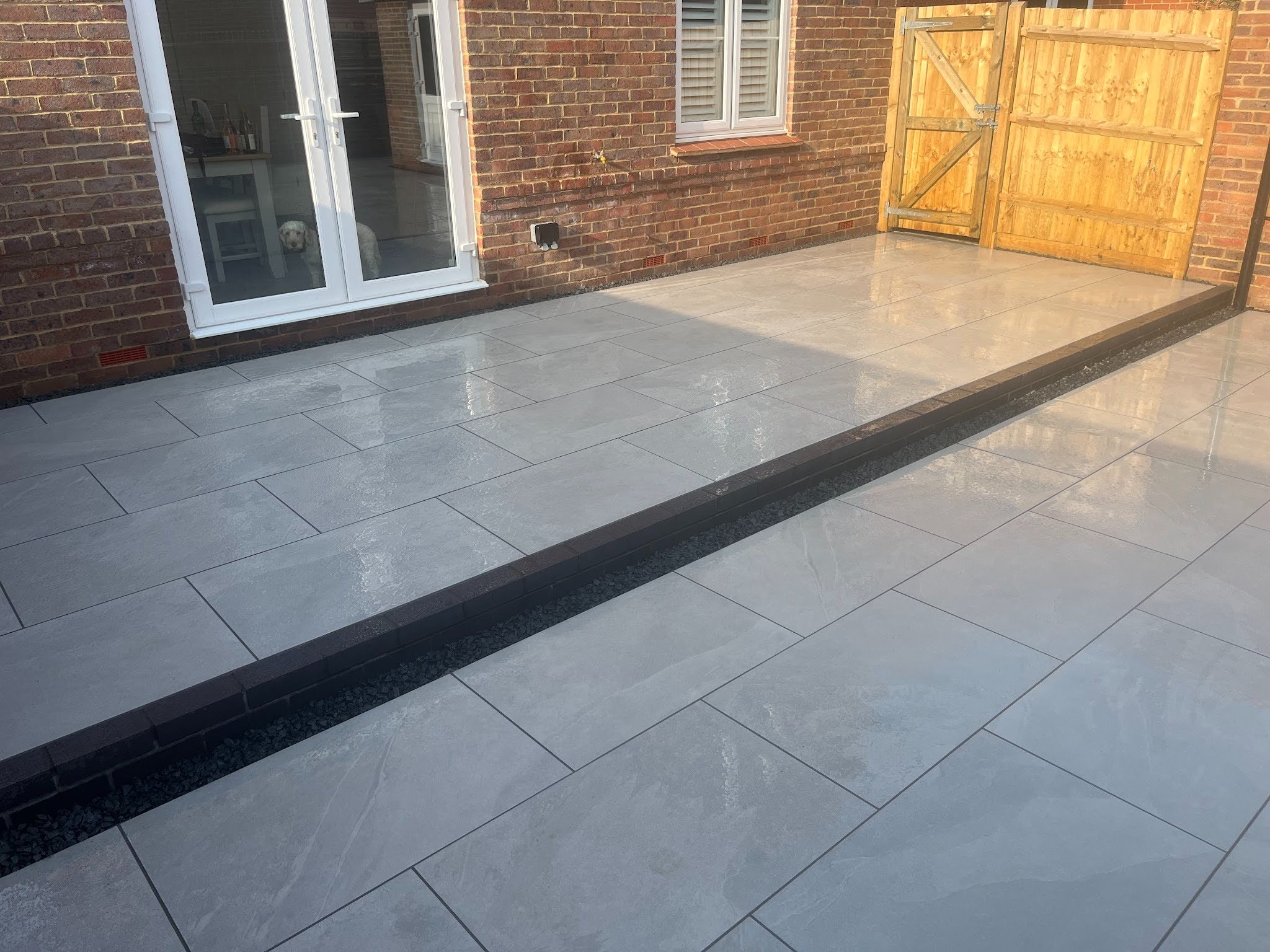The patio after a fresh wash &mdash; both terraces reading as a single resolved surface, the step riser anchoring the centre of the composition, French doors and side gate opening onto the upper tier.