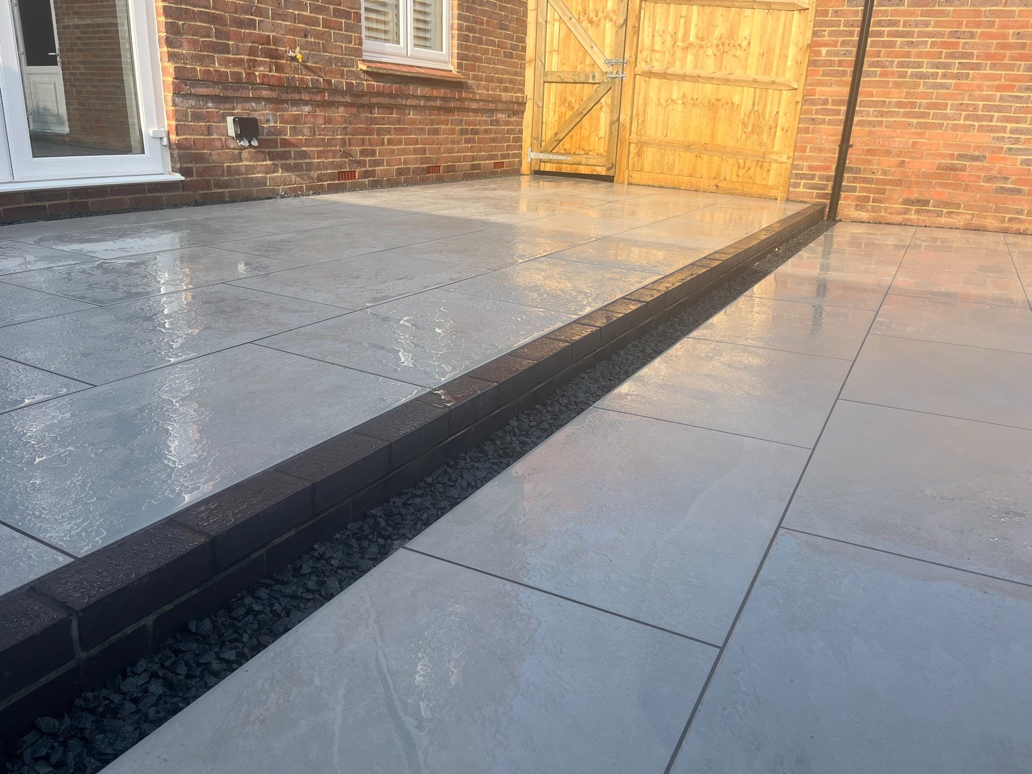 Detail of the step between the two terraces &mdash; solid blue engineering brick riser, Derbyshire Peak gravel channel at the base, carbon pointing reading as crisp dark lines between the Victoria Grey porcelain slabs. Photographed wet.