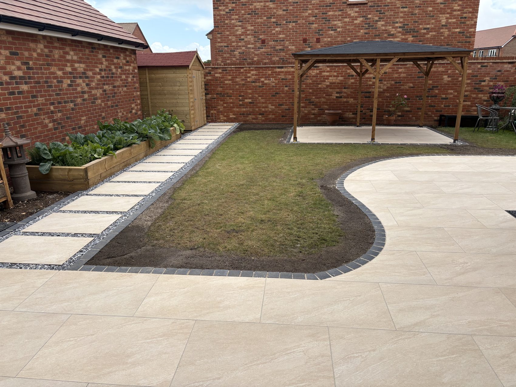 The finished Warsash garden: a curved Valour Beige porcelain terrace framed in black limestone, with a stepping-stone path, gazebo patio and raised vegetable beds beyond.