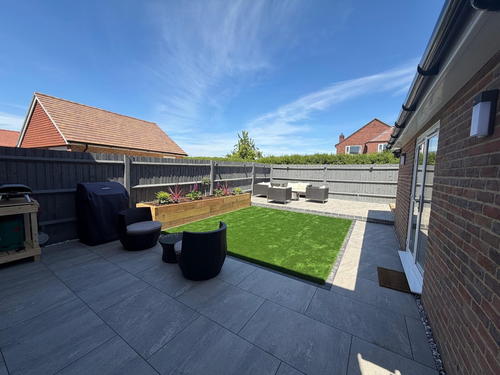 New-build garden in Warsash finished in large-format grey porcelain wrapping a central artificial lawn, with an oak sleeper raised bed and rattan seating, 2026