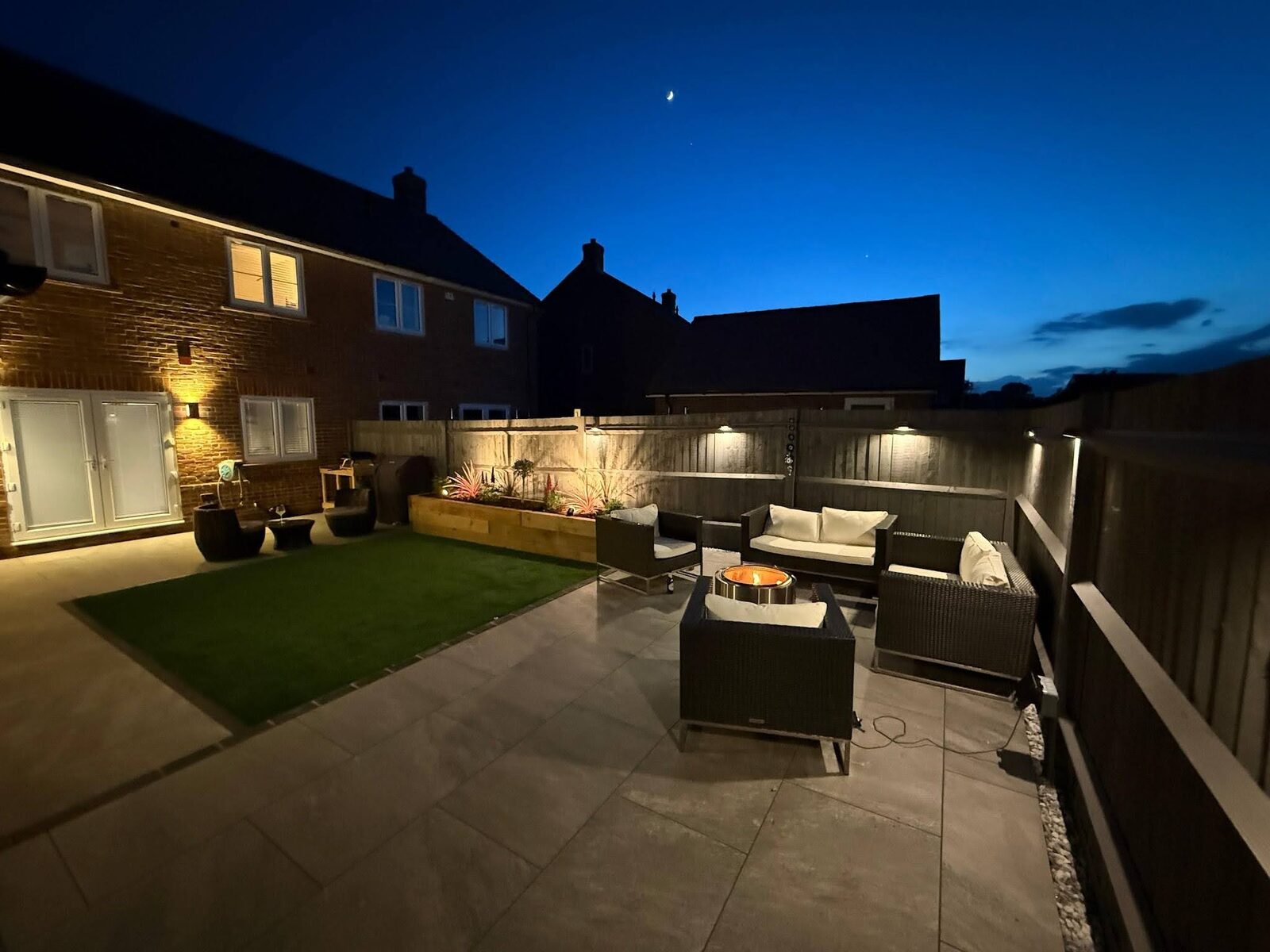 The rear terrace at dusk, lit for the evening with the seating area and framed lawn in view.