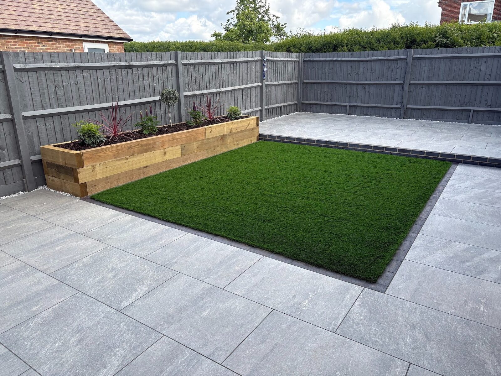 The oak sleeper raised bed running along the boundary, planted with phormium and clipped evergreens beside the framed lawn.