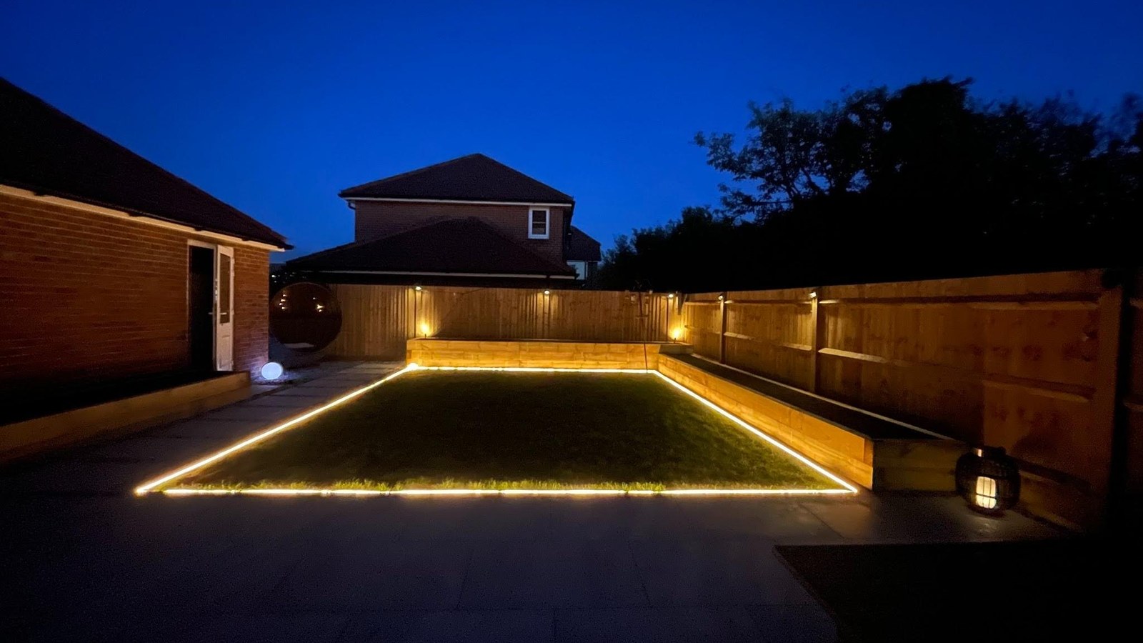 Garden composition at dusk — recessed LED strip framing the lawn perimeter, fence-panel uplighters, large-format porcelain terrace, Warsash