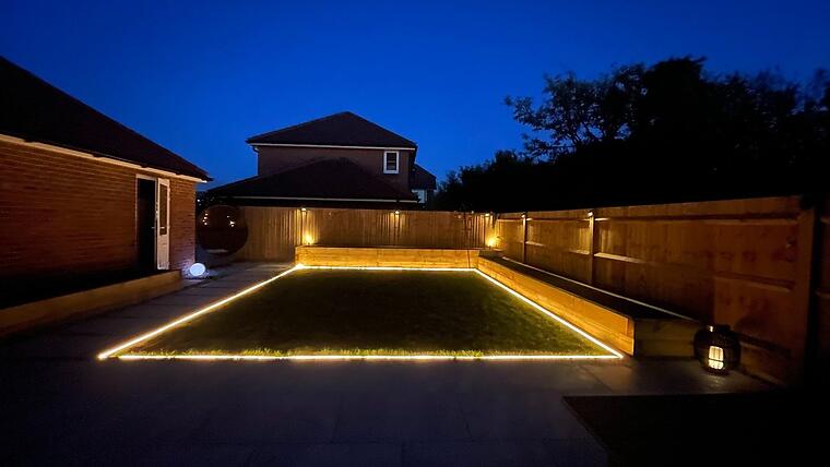 Garden composition, Warsash -- Garden at dusk — recessed LED lighting framing the lawn perimeter, fence-panel uplighters glowing, two-terrace porcelain hardscape.